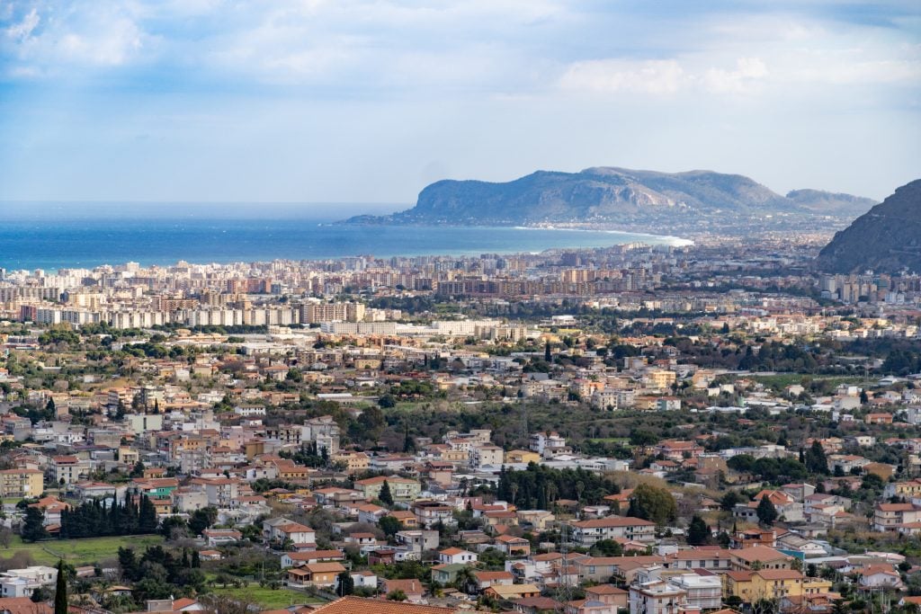 A long-distance view over the hills surrounding Palermo, covered with homes, mountains and ocean in the distance.