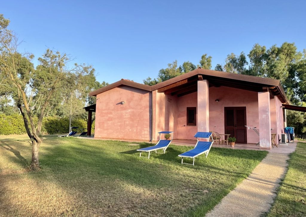 A pink house set on a big lawn, two blue lounge chairs in front of it.