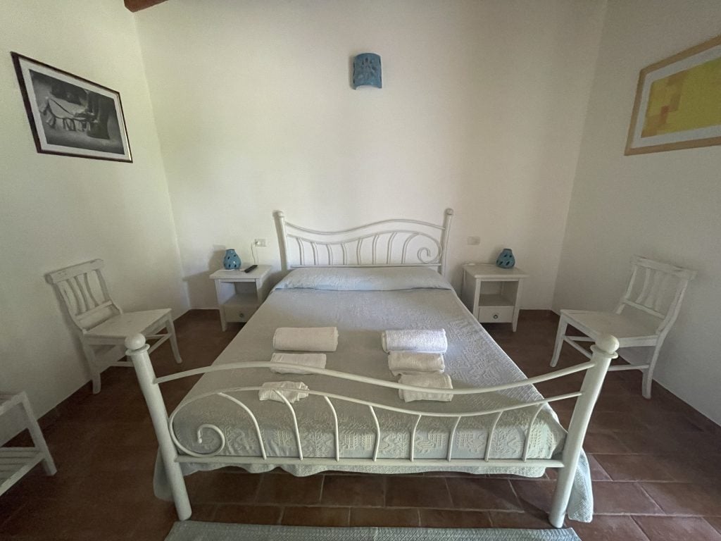 A simple room with a wrought-iron bed, two bedside tables and two basic wooden chairs surrounding it.