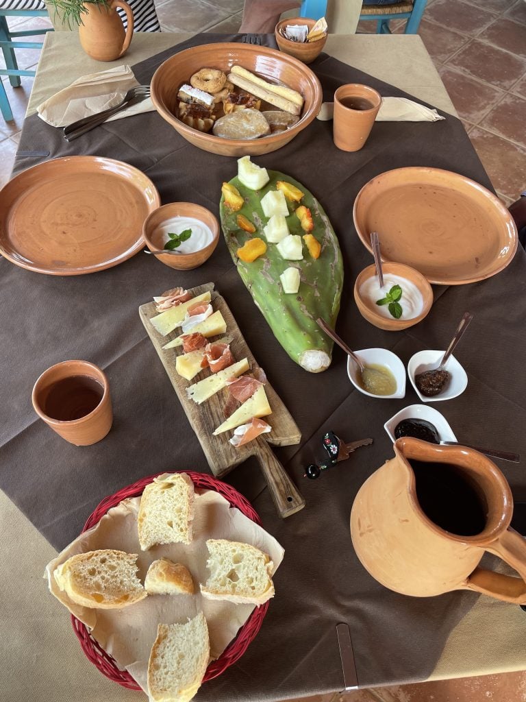 A breakfast spread with bread, cheese, meat, lots of fresh fruit, yogurt, and coffee.