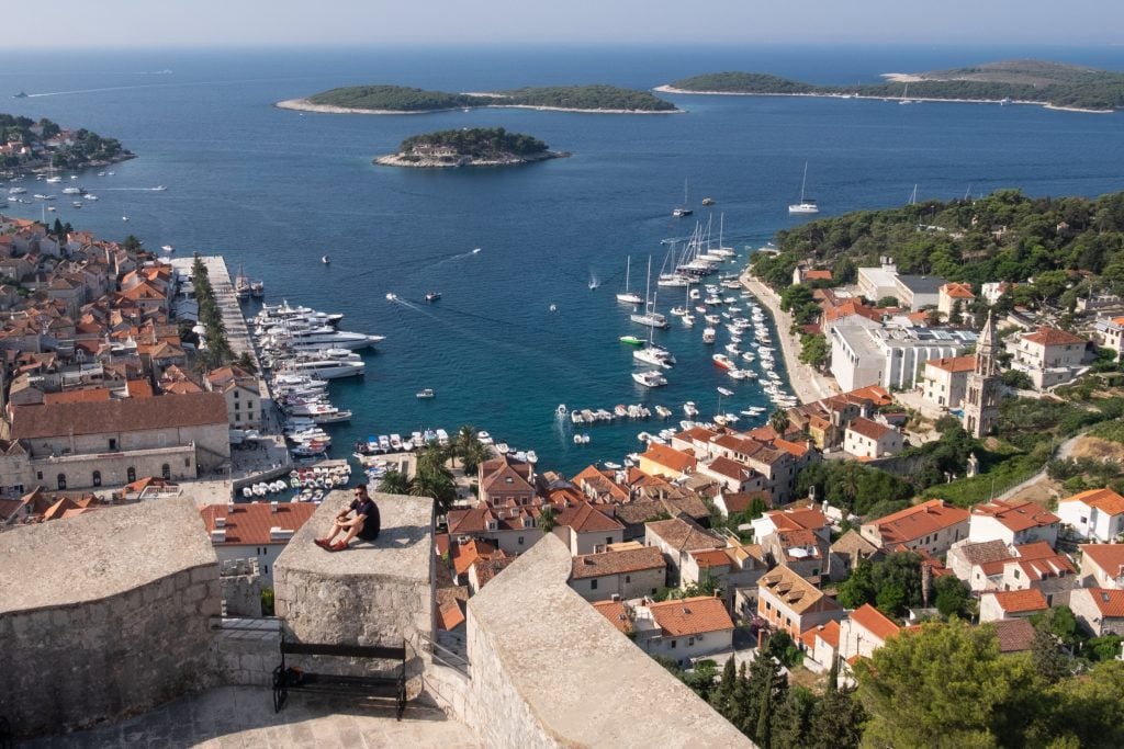 Charlie sitting on the edge of the stone fortress overlooking Hvar town with lots of orange roofs and the ocean, with several rocky green islands in the distance.