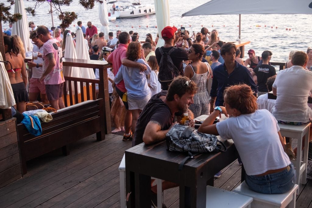A crowd of young people at an outdoor bar perched over the water.
