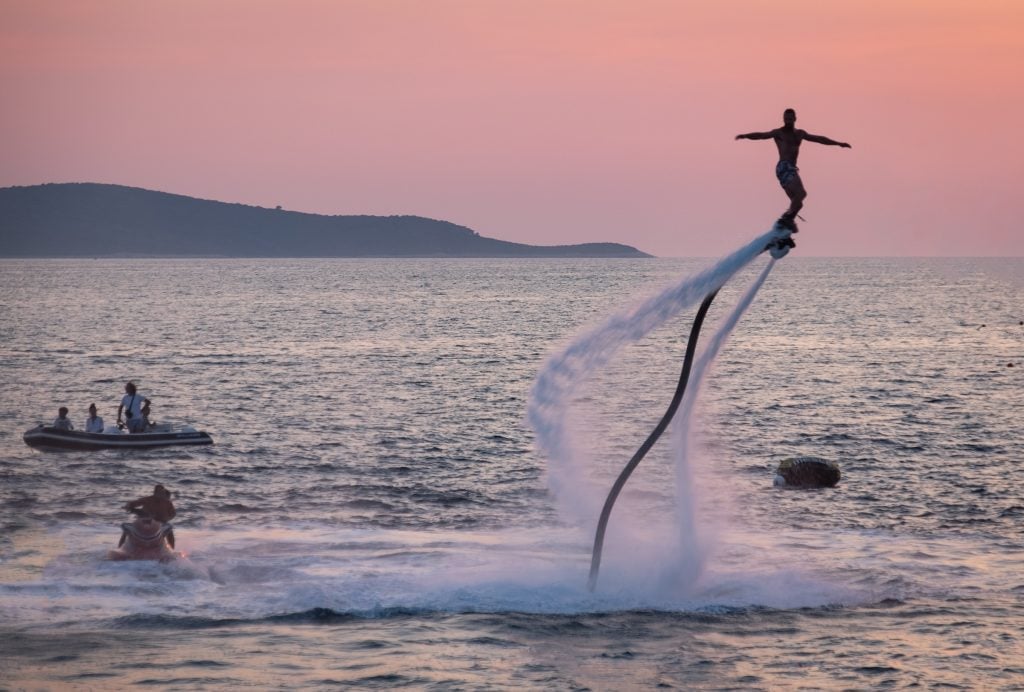 A man flying through the sky on one of those water thins that pushes water through the bottom of your feet and lets you soar through the sky.