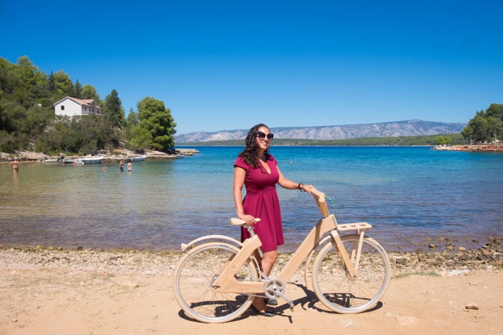 Kate standing in front of a beach in Hvar, holding a bicycle made of wood.