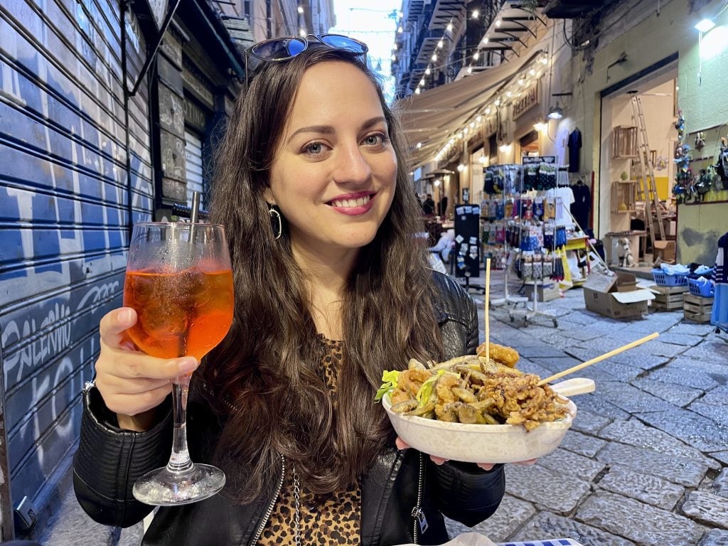 Kate smiling, sitting at an outdoor table, holding up an Aperol spritz in one hand and a bowl full of fried seafood in another.