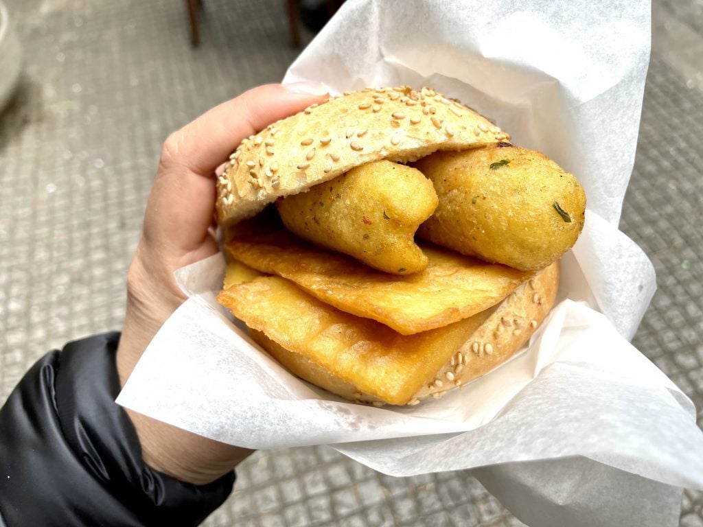 A sesame roll with flat chickpea fritters and potato croquettes in it.