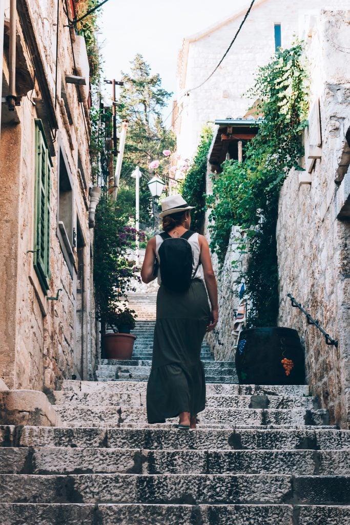 A woman walking up a stone staircase in Hvar wearing a long black dress and a Panama hat.