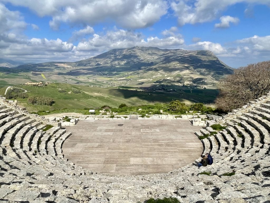 The view from the top back row of a half-moon shaped amphitheater, with views over a hilly green countryside and a blue sky studded with puffy clouds.