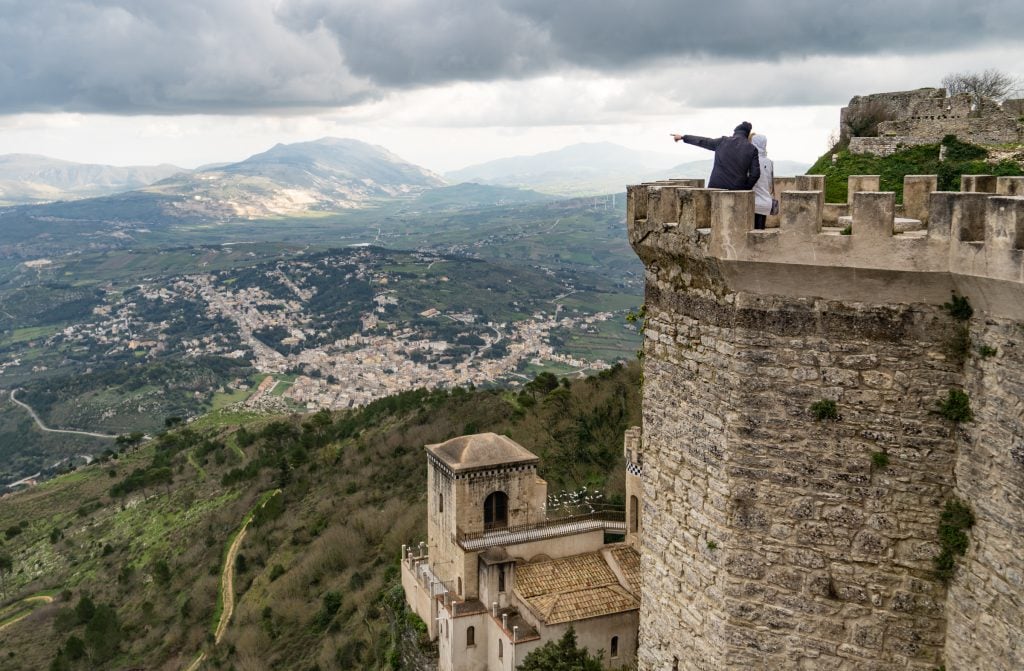 A couple standing in a medieval tower in Erice, Sicily, looking over the mountainous landscape underneath a cloudy sky.