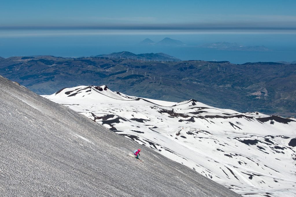 A skier skiing down shallow white snow on Mount Etna, the Aeolian Islands in the background.