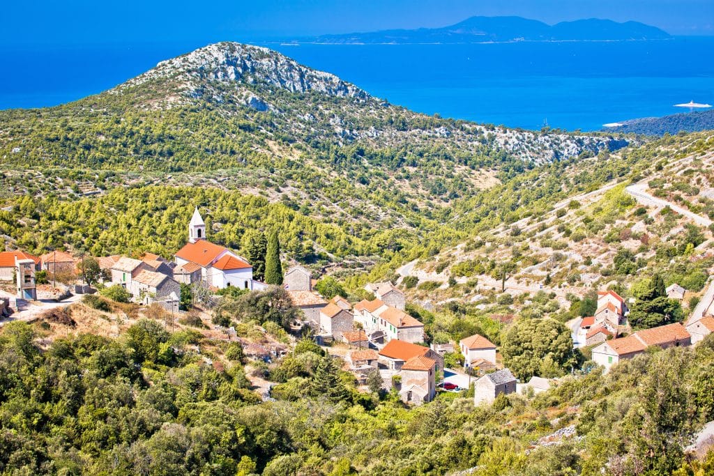 A tiny village of white buildings with orange roofs perched on a hill in Croatia, the sea not far behind in the distance.