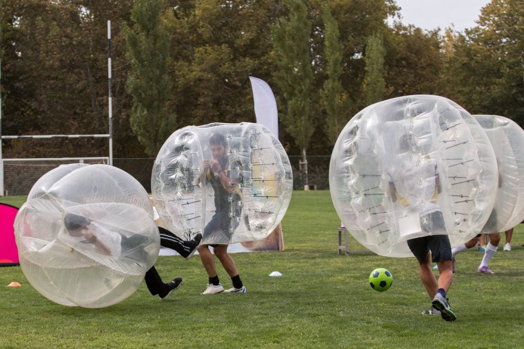 People running around wearing giant plastic balls around them, kicking soccer balls.