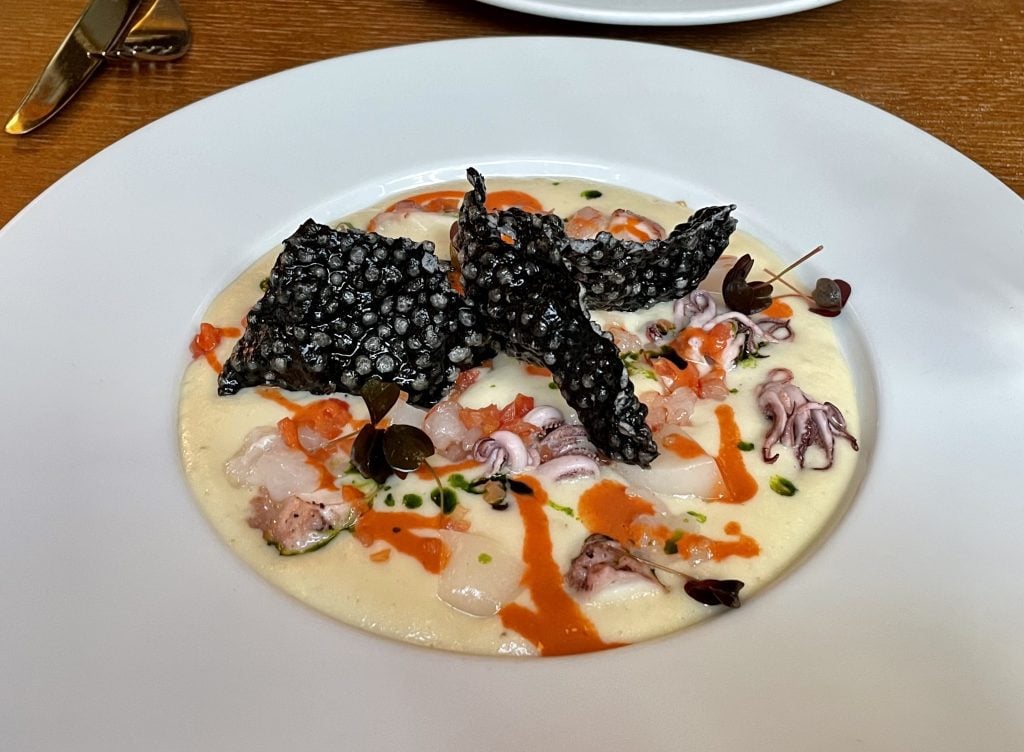 An appetizer that looks like various pieces of shellfish in a soup with a black cracker-like thing broken on top.