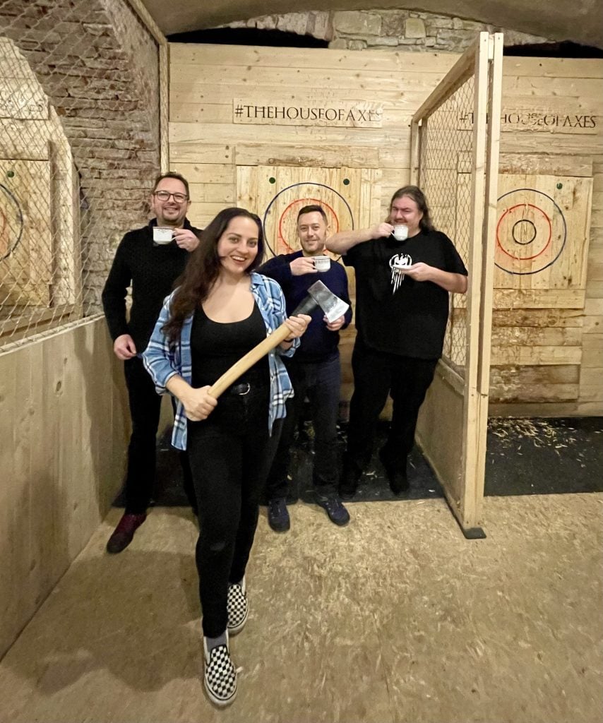 Kate holding an axe in front of a wooden target and standing in front of her three male friends who are all drinking from china teacups and laughing.