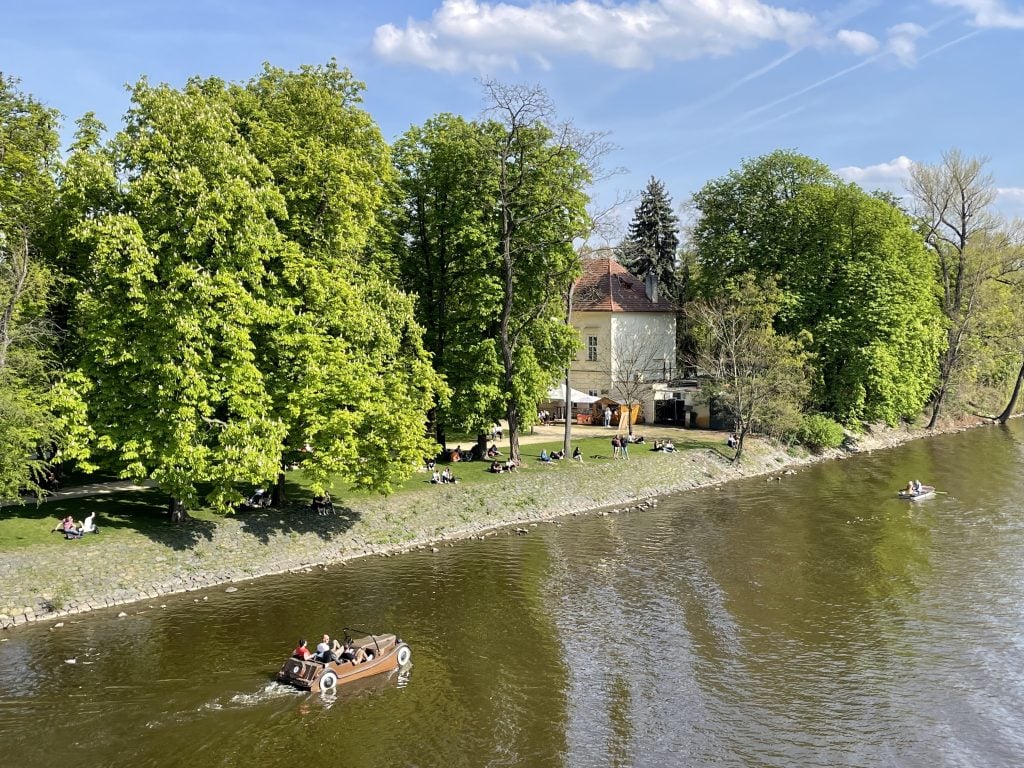 A boat shaped like an antique car pedaling past an island in the river.