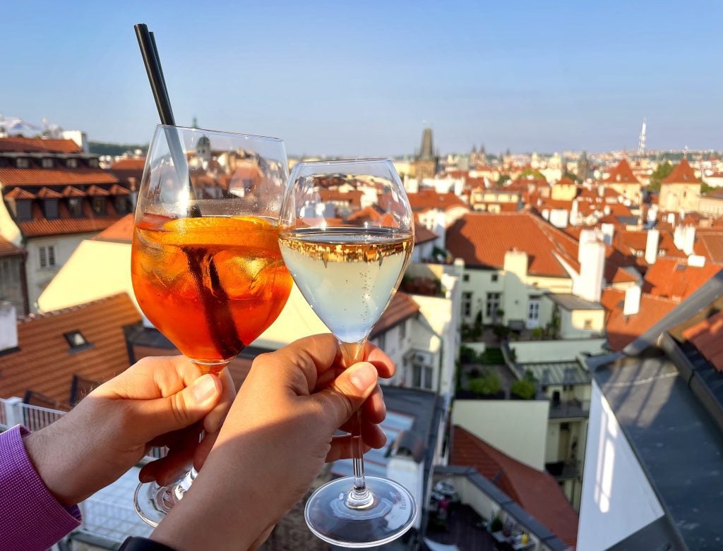 Two hands clinking an aperol spritz and a glass of champagne over a backdrop of orange roofs and blue sky.