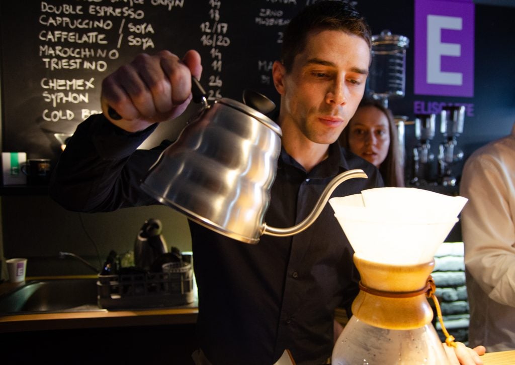 A man pouring hot water from a silver teapot into a Chemex glass coffee vial.