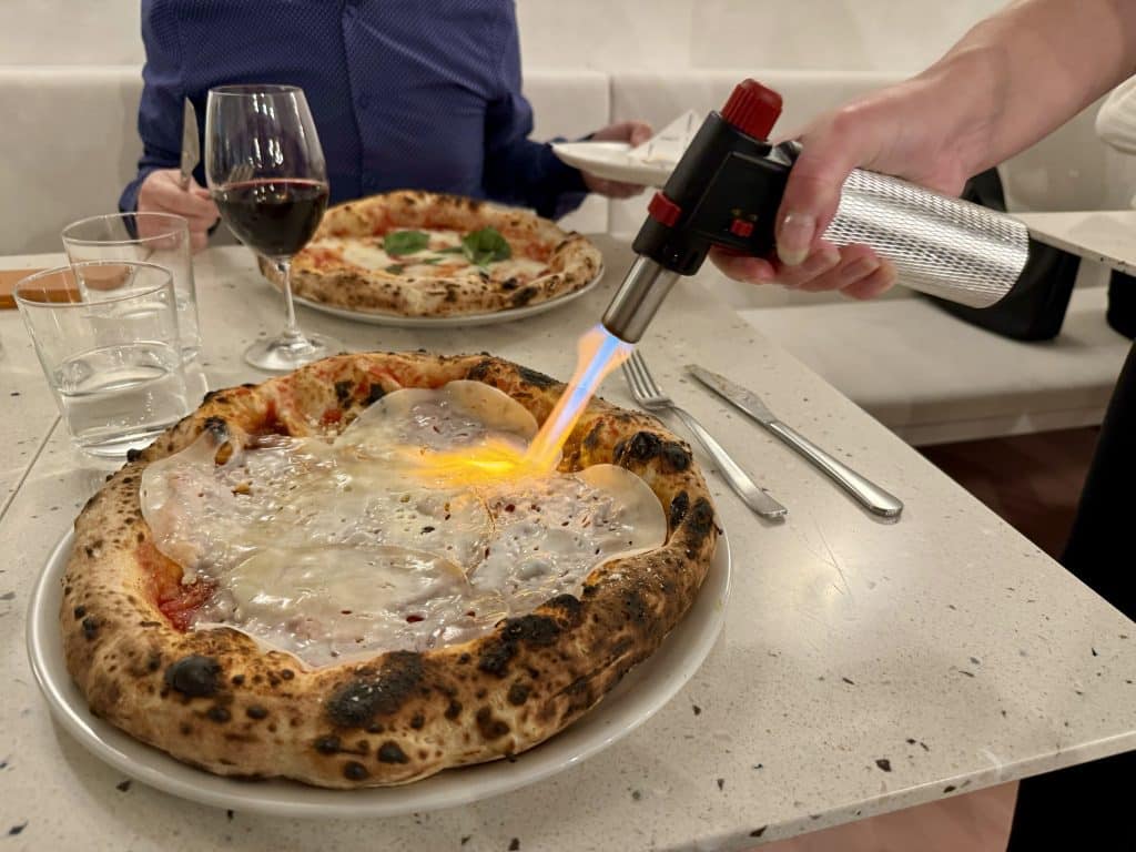 Two pizzas on a restaurant table -- as a server holds a flame-throwing torch and melts the cheese on one pizza.