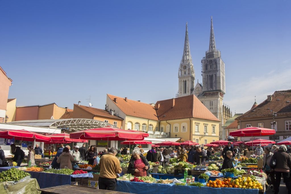 A fresh produce market outside in Zagreb with lots of red umbrellas, a church tower in the background.