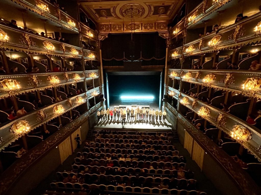 A theater with a cast taking their bows on stage, the sides of the theater filled with lit up box seats.