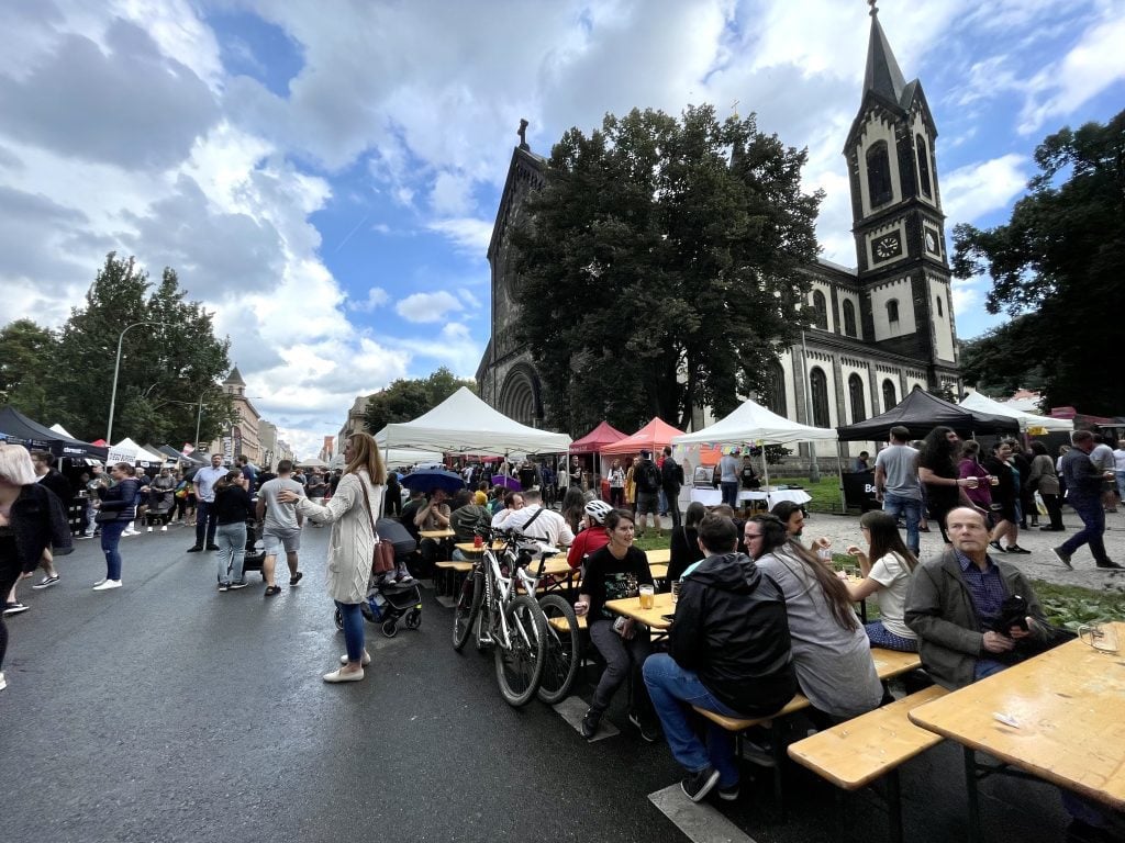 Groups of people sitting at wooden tables surrounded by food stalls in front of a Gothic church in Prague.