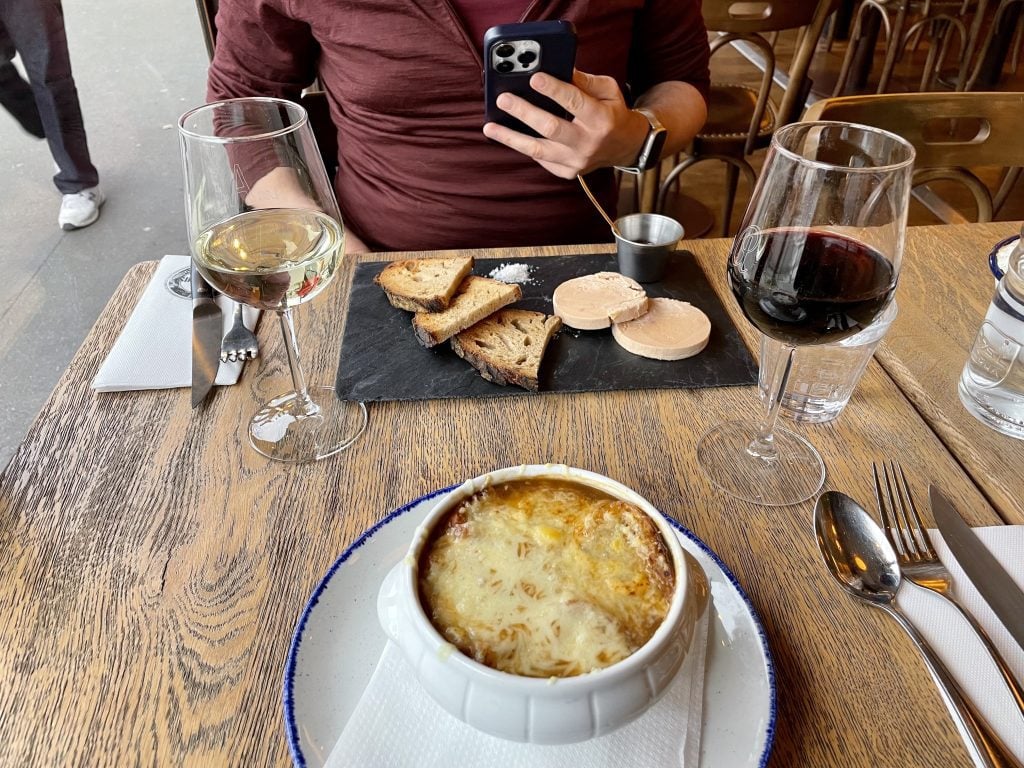 Sitting at a French cafe: a bowl of cheese-covered French onion soup and a glass of red wine, while across the table is toast points with foie gras and a glass of white wine.