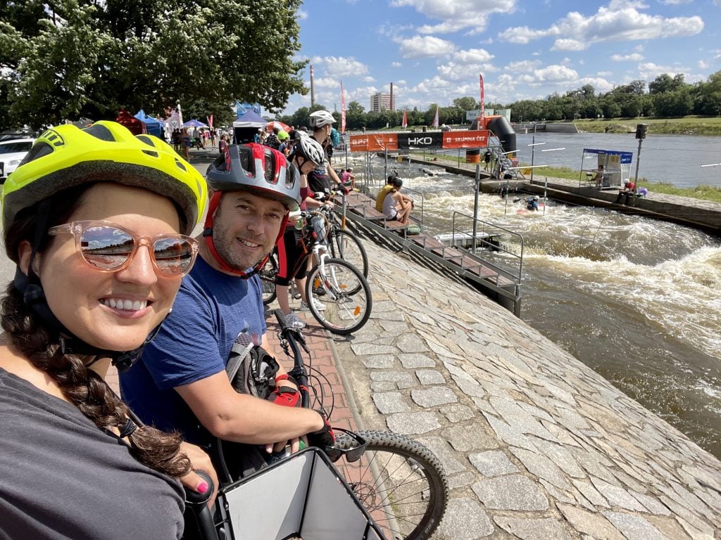Kate and Charlie taking smiling selfies with bike helmets on, standing atop their bikes, next to a river with people kayaking by.