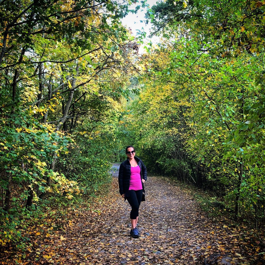 Kate stands on a forested path wearing a pink workout top, black pants and cardigan.