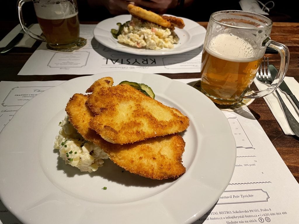 A plate with two thin pieces of chicken schnitzel on top of potato salad, next to a half-finished beer.