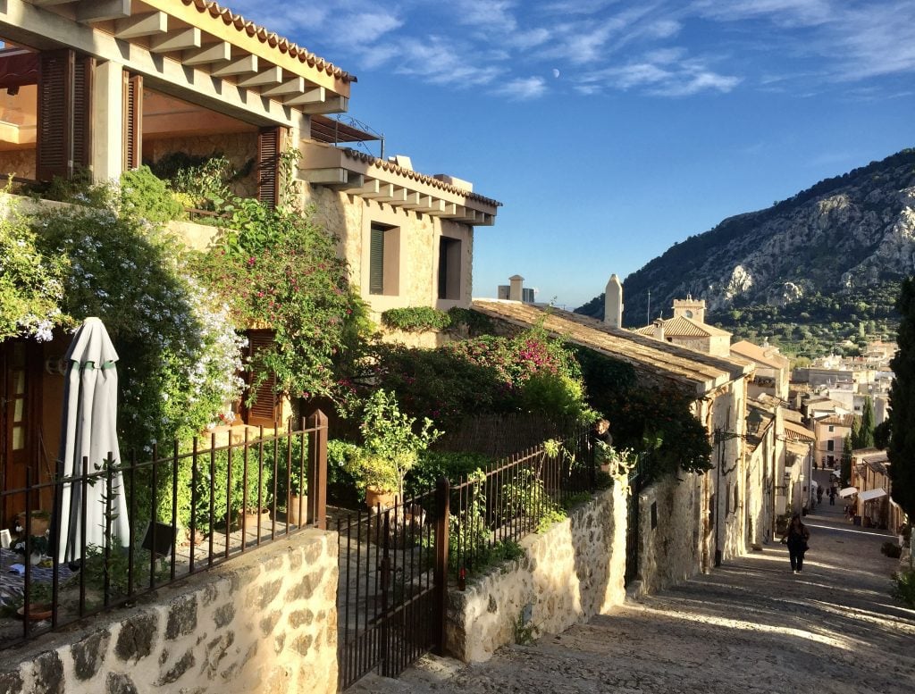 A cobblestone street with homes on the right and mountains and blue sky in the background in the town of Pollença on Mallorca