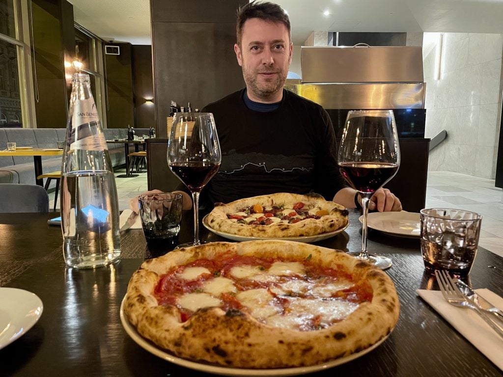 Two pizzas and two glasses of red wine on a table, Charlie sitting behind one of them and smiling.