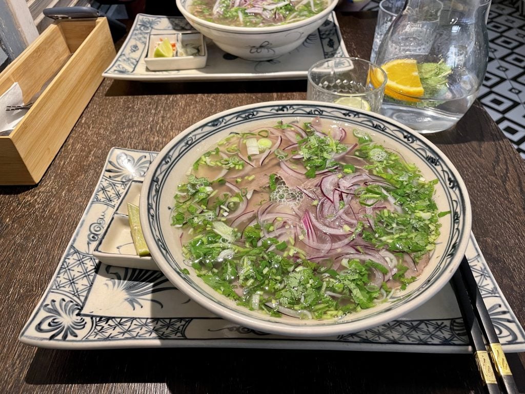 A rich bowl of beef pho topped with sliced onions and fresh herbs.