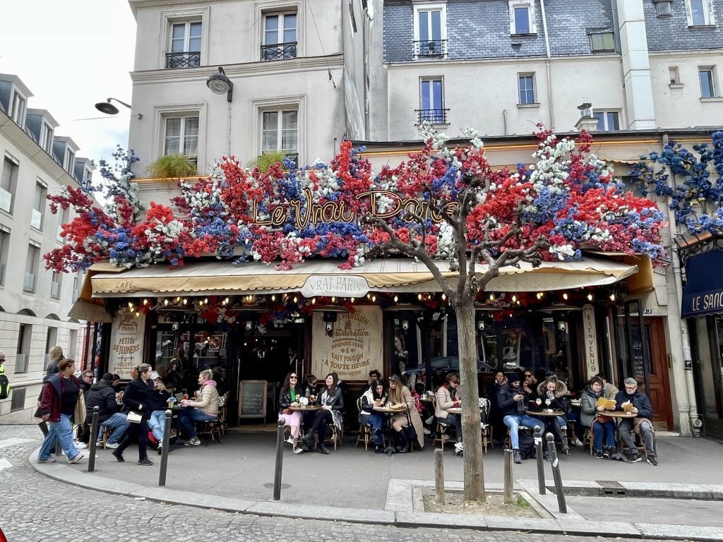 A Parisian cafe with an awning topped with red white and blue flowers.