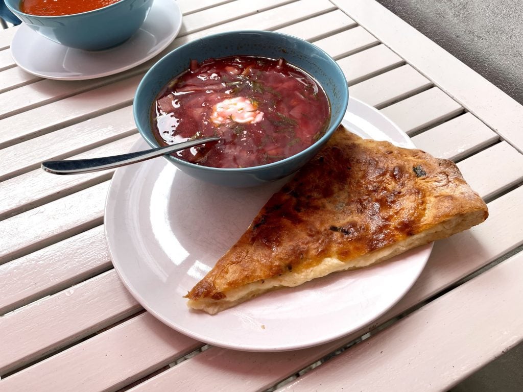 A bowl of deep red borscht topped with a dollop of sour cream, next to a slice of khachapuri, Georgian cheese-filled bread.