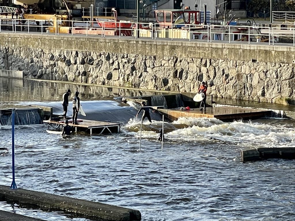 Five surfers taking turns jumping onto an artificial wave in the middle of the river.