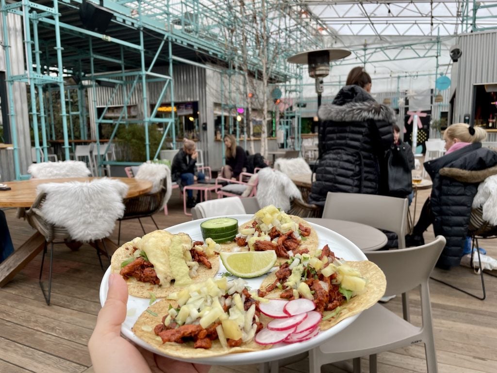 A hand holding a plate of tacos al pastor with pork and pineapple in a big market area.