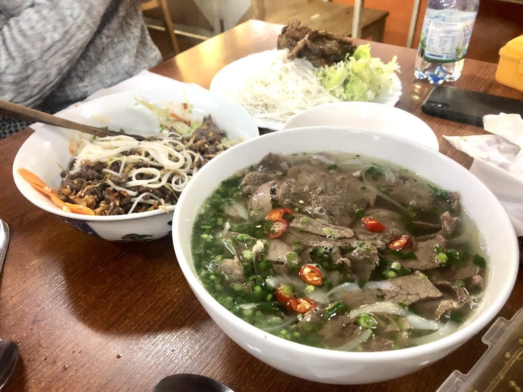 A bowl of Vietnamese pho next to a bowl of noodle and a plate of roasted pork and rice.