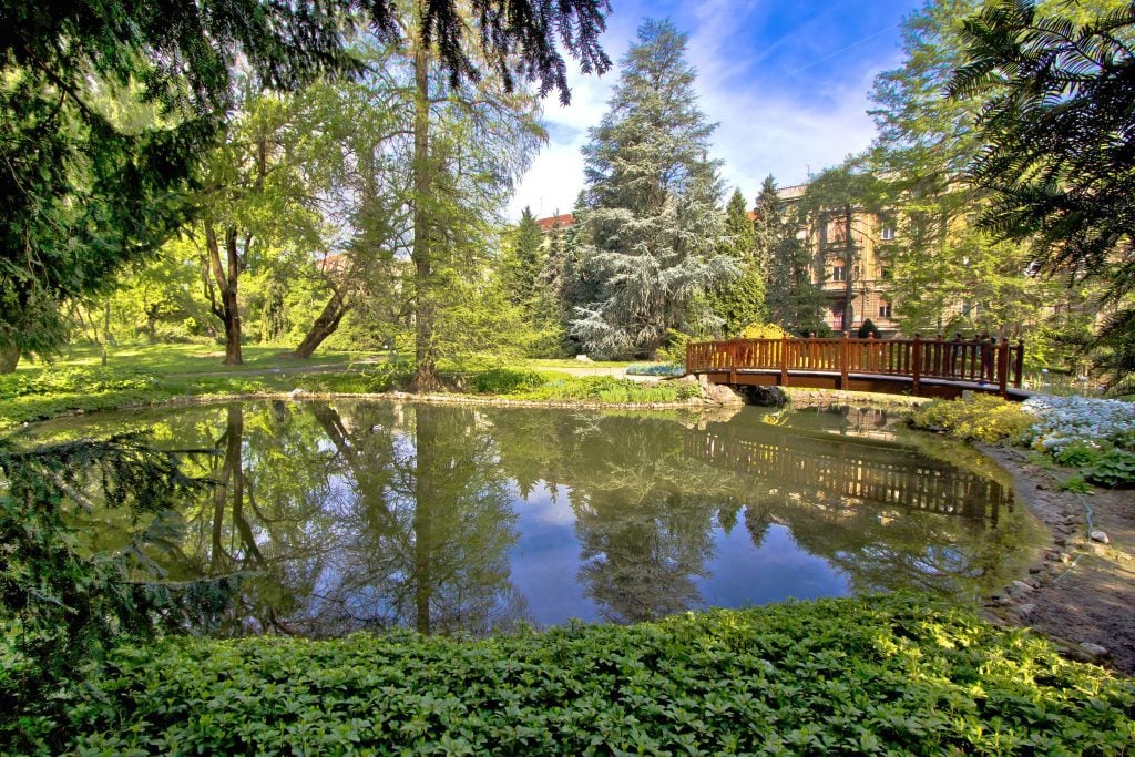 A red wooden. bridge crossing a small pond surrounded by trees in the Zagreb Botanical Garden.