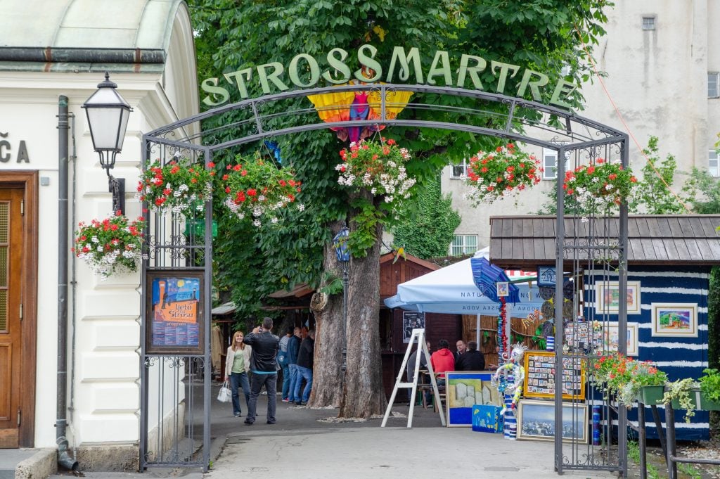 A gate reading "Strossmartre" in front of a narrow street lined with trees and cafes.