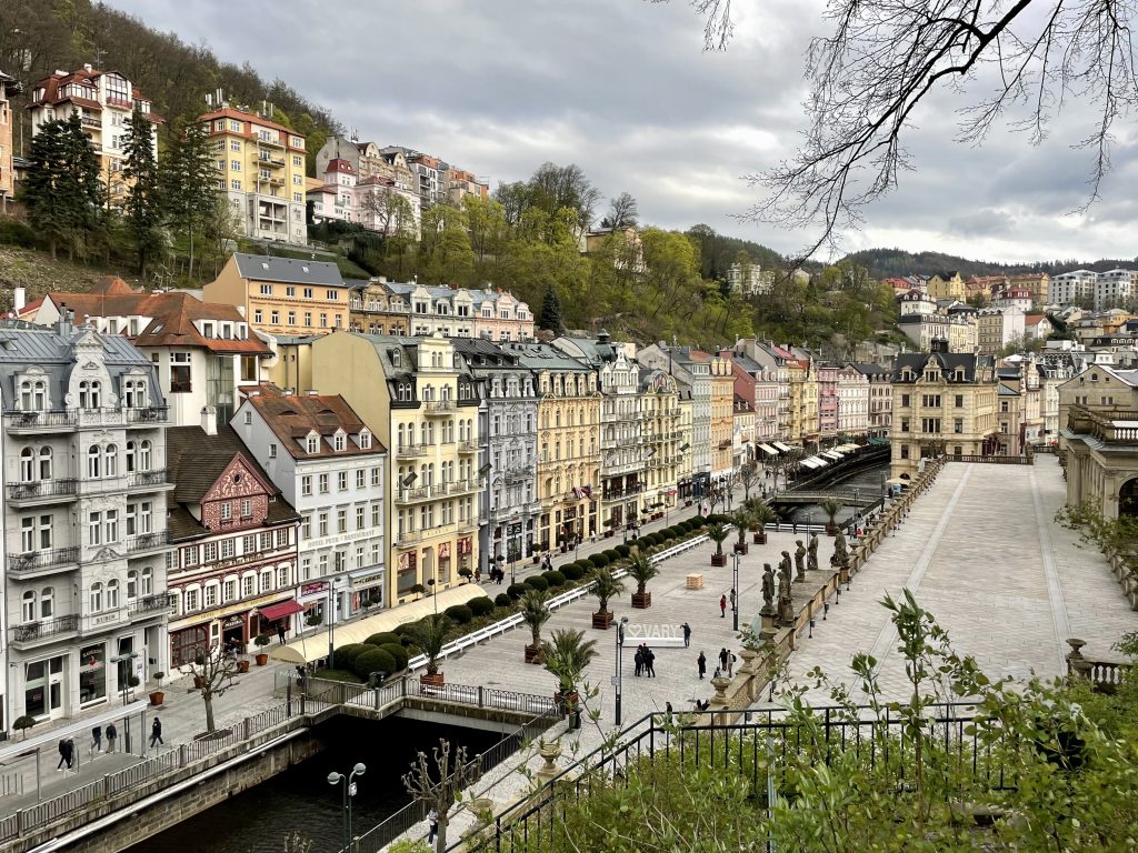 The city of Karlovy Vary, with tall crenellated buildings in pale colors lining a calm river, people walking alongside the river. Surrounded by hills in each direction.