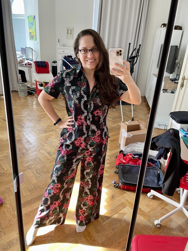 Kate posing in a mirror wearing a short-sleeved button-up jumpsuit with flared legs. It's navy blue with a pattern of pink roses, green leaves, and white flower chains.