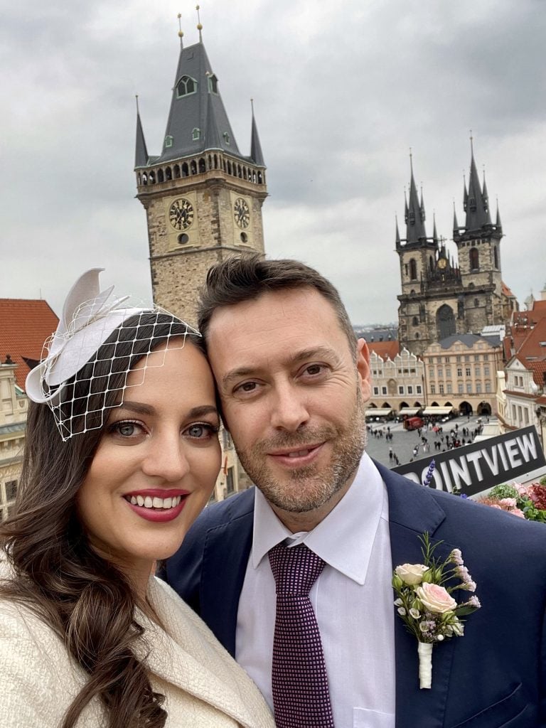 Kate and Charlie posing over the skyline view of Prague, gothic churches in the background. Kate wears a white fascinator and suit; Charlie wears a navy suit with a purple tie and a boutonniere.