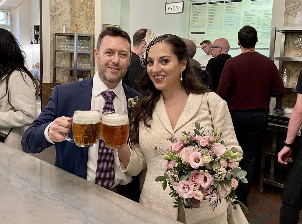 Charlie and Kate stand in a busy pub, clinking pints of beer. They wear wedding outfits (Charlie in a navy suit and purple tie, and Kate in an ivory skirt suit and fascinator) and Kate carries a bouquet of pale pink and purple flowers.