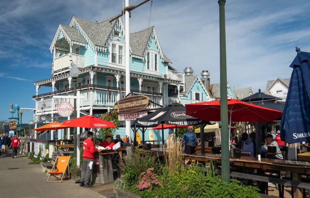 A crowd of people enjoying drinks at an outdoor bar underneath umbrellas, next to a tall pale blue Victorian house with a wraparound porch.