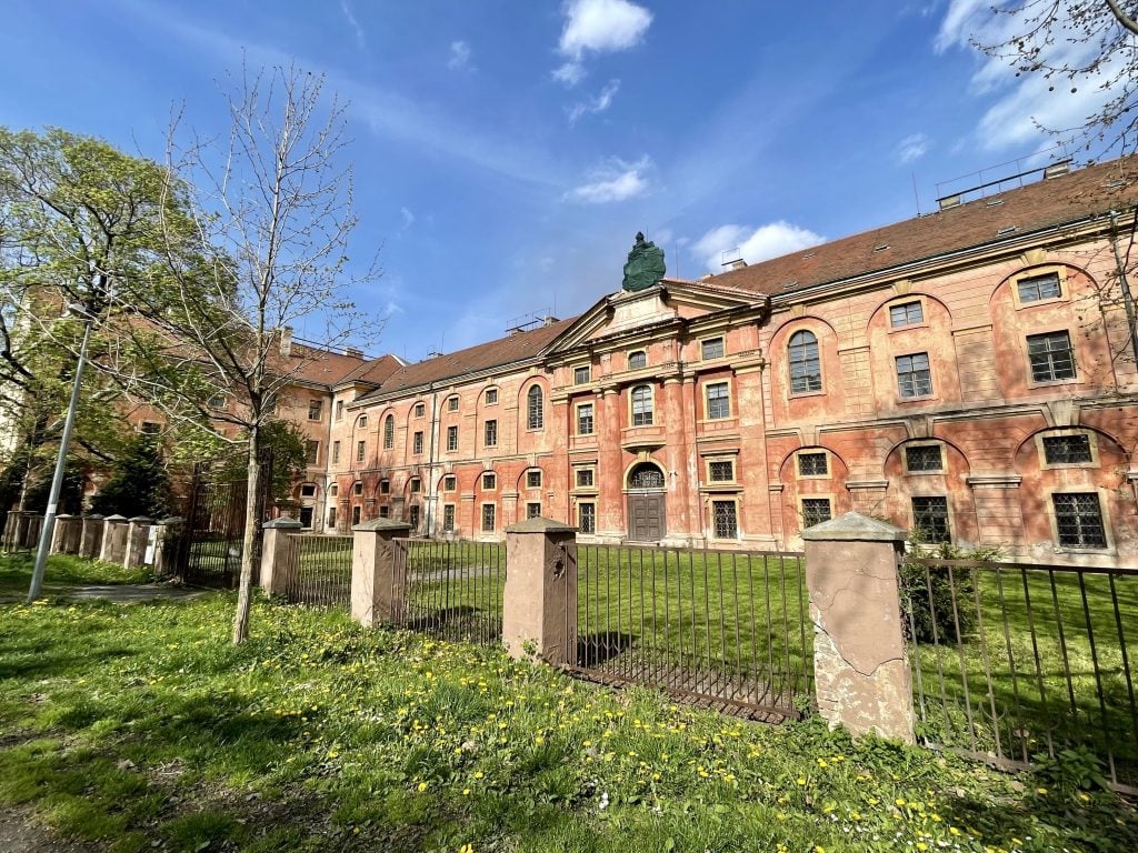 A red and yellow Bohemian building underneath a blue sky, surrounded by an overgrown lawn in the spring.