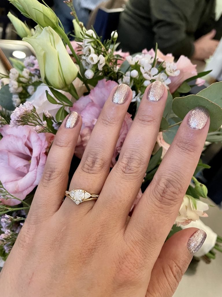 Kate's hand with a gold engagement ring next to a thin curved band with three small diamonds. She has a pale pink metallic manicure.