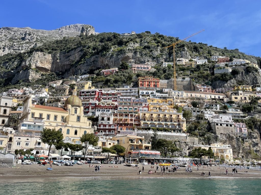 The town of Positano on the Amalfi Coast, with steep hills stacked with small homes, leading to a small gray beach and the teal ocean.