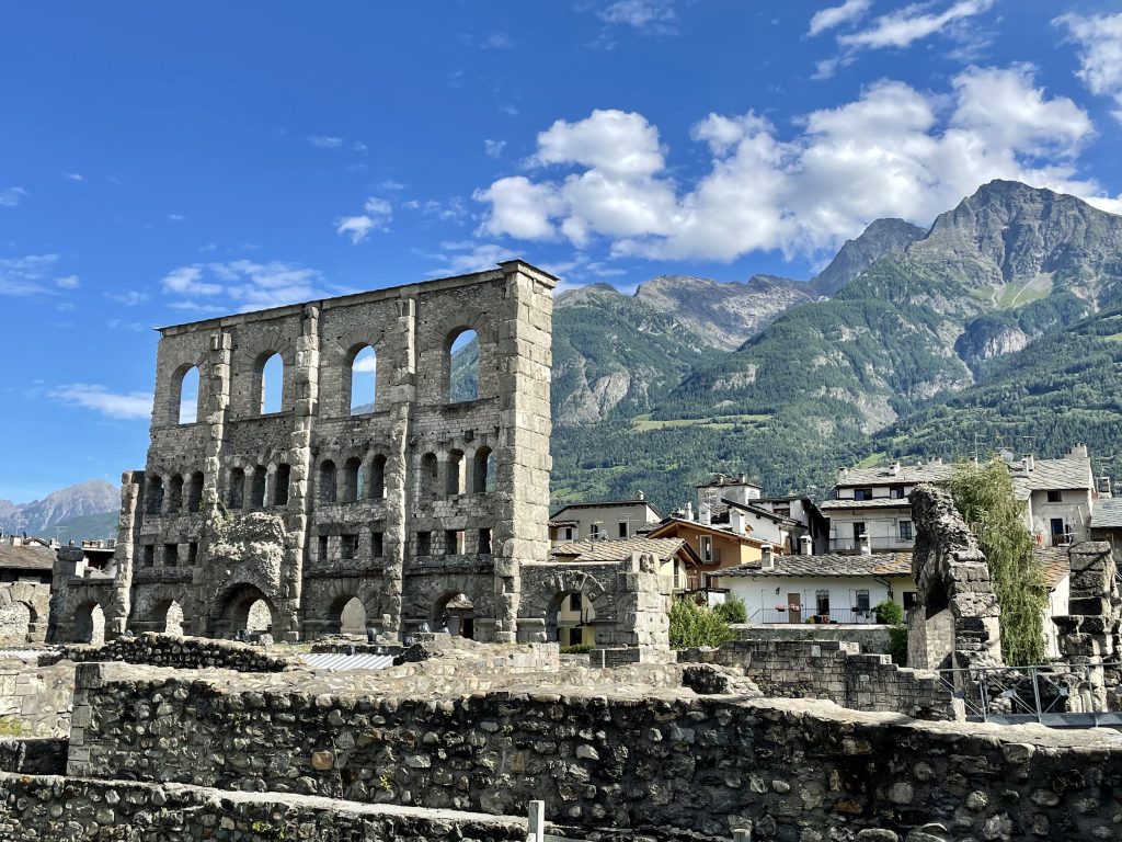 Roman ruins of an amphitheater in the foreground, and rows and rows of Italian homes followed by tall craggy green mountains in Aosta, Italy.