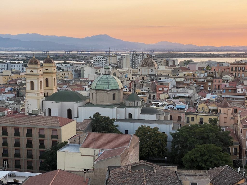 A sunset view over the city of Cagliari, with lots of views of rooftops and green-domed churches, mountains in the background.