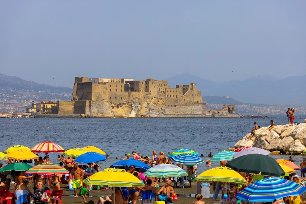 A tall stone castle on the edge of the sea in Naples, a beach with lots of colorful umbrellas in the foreground.